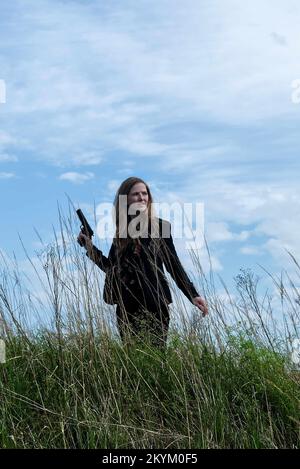 donna in tuta con pistola in piedi nel log grass. stile copertina libro a distanza media. Foto Stock