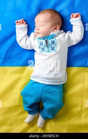 Rivne, Ucraina 25 aprile 2022: Un neonato vestito nazionale si trova e dorme sulla bandiera dell'Ucraina, bambini nati durante la guerra. Foto Stock