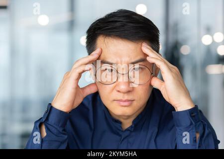 Primo piano foto di pensare triste uomo d'affari asiatico, uomo in occhiali guardando la macchina fotografica deluso, capo in camicia all'interno dell'ufficio al lavoro Foto Stock