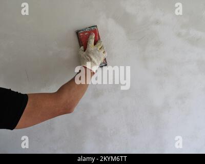 sbucciando irregolarità su una parete di stucco, intonacando una superficie di parete intonacata di fresco con smeriglio, un uomo in nero lavora a sbucciare una parete Foto Stock