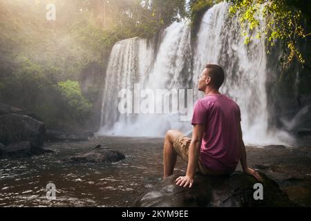 Uomo che riposa sulla pietra di fronte ad alta cascata in montagna in paesaggio tropicale a Camoboda Foto Stock