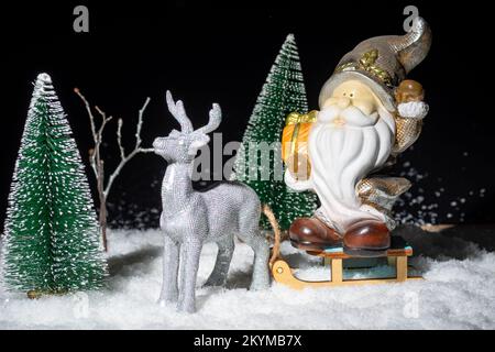 Biglietto di Natale con una renna che porta una slitta con Babbo Natale con regali nella neve tra gli abeti di notte, creativo. Storia di Natale. Nuovo Foto Stock