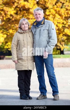 Coppia anziana in piedi insieme indossando giacca, albero giallo autunno, guardando la macchina fotografica, ritratto a tutta lunghezza Foto Stock