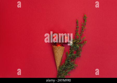 Tre giocattoli rotondi nel cono del waffle si trovano come gelato delizioso e accanto ad esso è ramo verde dell'albero di Natale su fondo rosso. Concetto di celebrare le fredde vacanze invernali in caldo. Vista dall'alto, disposizione piatta Foto Stock