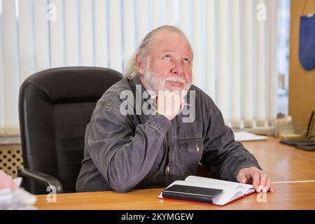 Un uomo anziano guarda in su pensieroso, un pensionato al posto di lavoro in una sedia Foto Stock