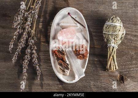 Vassoio porta incenso in ceramica bianca con pietre in cristallo minerale e asta di Aroma. Fascio di salvia salvia e lavanda sullo sfondo rustico di legno Foto Stock