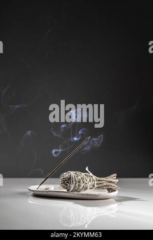 Burning Aromatic incense Smoky Stick e Salvia Sage su vassoio bianco per meditazione e relax su sfondo minimalista scuro. Aromatherapy Smoke è in uso Foto Stock