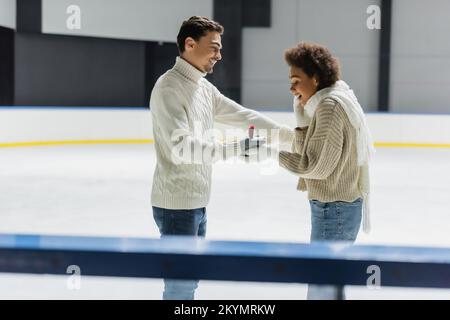 Vista laterale di un uomo sorridente che mostra l'anello di fidanzamento alla ragazza afroamericana sulla pista di pattinaggio Foto Stock