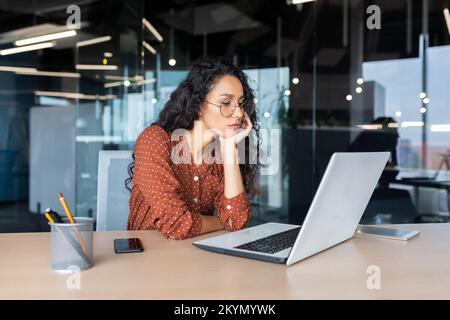 Triste grumpy latinoamericana che lavora all'interno dell'ufficio, donna d'affari è annoiato guardando lo schermo del laptop. Foto Stock