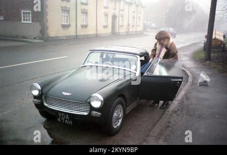 1980s, un primo scatto di mattina e una giovane donna che indossa una giacca di pelle che lotta per ottenere borse di macchine fotografiche in una piccola macchina sportiva Austin-Healey Sprite 2 porte, parcheggiata in una layby di una strada, Inghilterra, Regno Unito. Sul marciapiede si trova una borsa con lente per fotocamera. In produzione dal 1958 al 1971, la Sprite è stata progettata dalla Donald Healey Motor Company e prodotta presso la FABBRICA MG di Abingdon, Oxford, Inghilterra. MG aveva la propria piccola auto sportiva a 2 porte, la Midget, praticamente identica alla Austin-Healey Sprite, ma con un distintivo diverso. Foto Stock