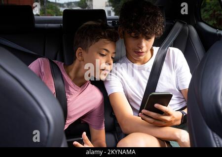 Fratelli che indossano le cinture di sicurezza che condividono lo smartphone mentre sono seduti in auto Foto Stock