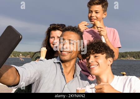 Uomo felice che prende selfie con la famiglia attraverso lo smartphone mentre si gode il gelato durante le vacanze Foto Stock