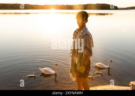 Ritratto di ragazzo felice in piedi vicino al lago con cigni che nuotano sullo sfondo Foto Stock