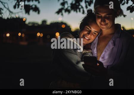 Donna sorridente che abbraccia un amico non binario utilizzando il telefono cellulare di notte Foto Stock