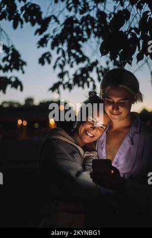 Sorridente persona non binaria che utilizza il telefono cellulare da un amico di notte Foto Stock