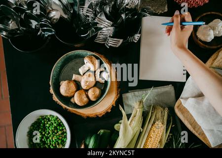 Mano di cuoco femmina scrivere ricetta in agenda da verdure e posate sul banco Foto Stock