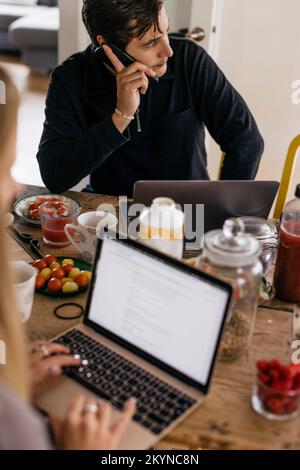 Freelance maschile che parla sullo smartphone mentre si siede con la colazione sul tavolo a casa Foto Stock