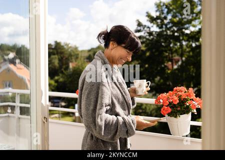 Vista laterale di una donna felice che tiene la tazza con uno smartphone mentre si trova in balcone Foto Stock