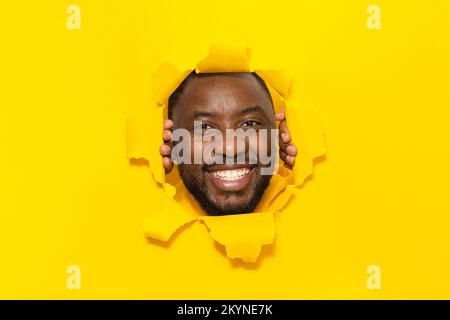 Felice uomo afroamericano che posa in carta gialla strappata, sorridendo guardando la macchina fotografica attraverso il foro sullo sfondo dello studio Foto Stock