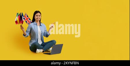 Sorridente studentessa di signora giapponese millenaria tiene molte bandiere, punta il dito in uno spazio vuoto, siediti a terra con un computer portatile Foto Stock