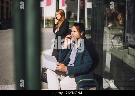 Imprenditore maschio che parla su smartphone mentre si siede con il computer portatile alla fermata dell'autobus durante la giornata di sole Foto Stock