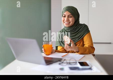 Giovane donna musulmana che mangia spuntini davanti al notebook a casa Foto Stock
