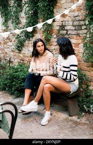 Amici femminili che parlano l'un l'altro mentre si siedono sulla panca vicino al muro di mattoni al partito del giardino Foto Stock