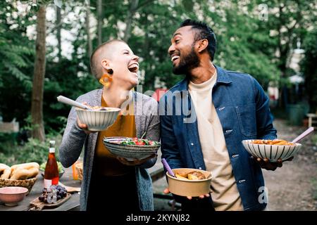 Amici maschili e femminili multirazziali ridendo e guardandosi mentre tengono le ciotole del cibo Foto Stock