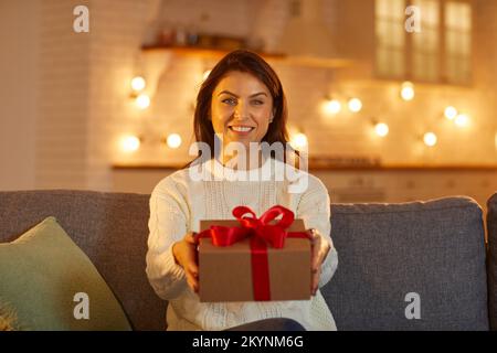 Donna felice guardando la macchina fotografica, sorridendo e dandovi la scatola con il regalo di Natale Foto Stock