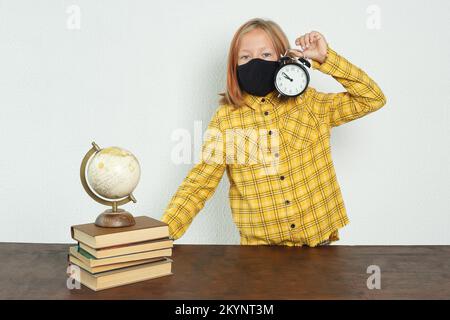 Concetto di istruzione. Una ragazza in una maschera si trova vicino al tavolo e tiene in mano una sveglia. C'è un globo e libri sul tavolo Foto Stock