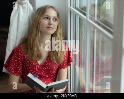 Una giovane ragazza in un vestito rosso siede sul pavimento vicino alla finestra con un libro nelle sue mani e guarda fuori dalla finestra. Foto Stock