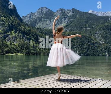 Una giovane ballerina danza su un palco in legno sul lago sullo sfondo delle montagne alpine Foto Stock