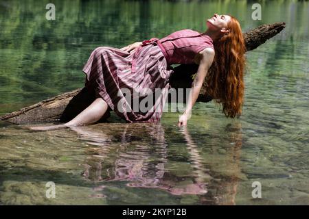Una giovane donna giace su un tronco con la mano in un lago di montagna color smeraldo Foto Stock