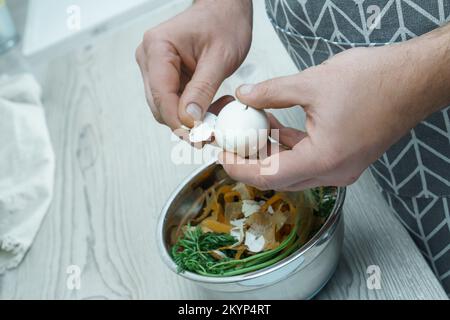 Mani umane che sbucciano il guscio di uovo bollito sopra ciotola di pelature vegetali. Persona cucinare sul tavolo da cucina, preparare gli ingredienti alimentari. Nutrizione sana, pr Foto Stock