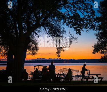 Silhouette di persone e alberi e barche a riva del lago con un tramonto arancione in lontananza al crepuscolo. Foto Stock