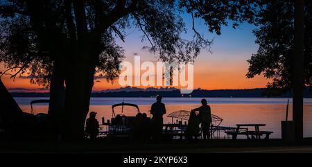 Silhouette di persone e alberi e barche a riva del lago con un tramonto arancione in lontananza al crepuscolo. Foto Stock