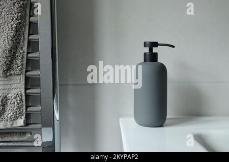 Dispenser di sapone grigio sul lavandino in un bagno rivestito di piastrelle grigie. Un asciugamano pende da un radiatore cromato Foto Stock