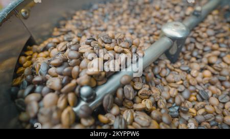 Primo piano dei chicchi di caffè mescolati e tostati. Azione. La macchina speciale mescola i chicchi di caffè per la lavorazione termica. Tostatura del caffè in macchina. Foto Stock