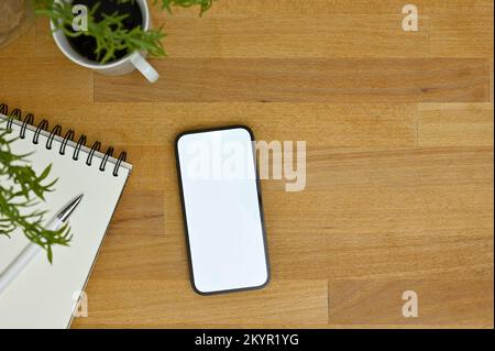 Vista dall'alto, Uno smartphone con schermo bianco mockup con blocco note a spirale e tazza da caffè su tavolo in legno. Foto Stock