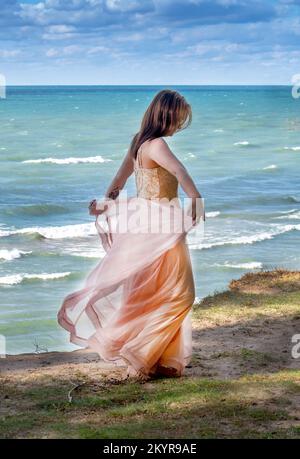 bella giovane ragazza in un abito rosa scorrevole, cammina lungo un bordo alto sopra un lago verde blu Foto Stock