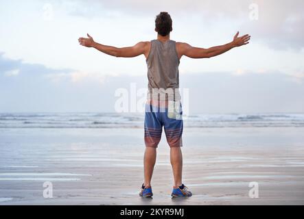 Abbracciando una bella mattina. Ripresa da dietro di un giovane uomo in piedi sulla spiaggia con le braccia sollevate verso il cielo. Foto Stock