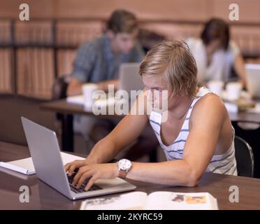 Fare ricerca per il mio saggio successivo. un giovane studente universitario che usa un computer portatile mentre studia in una biblioteca. Foto Stock