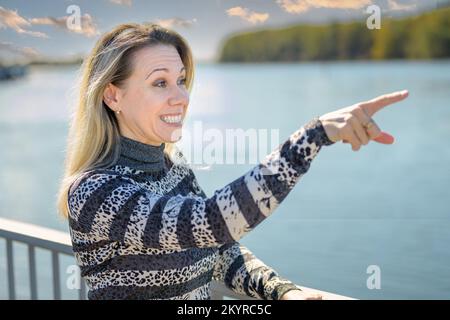 Vista laterale di una bella donna divertente con un sorriso luminoso che punta a qualcosa in lontananza con il dito su una ringhiera di fronte a un lago o di un'insenatura Foto Stock