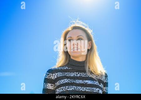 Basso angolo girato contro il sole di una donna di mezza età con lunghi capelli biondi contro un cielo blu in autunno Foto Stock