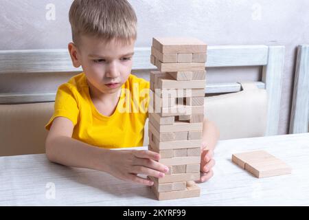 Interessato ragazzo biondo felice giocare gioco di bordo prendendo mattoni da torre di legno mantenere equilibrio avendo divertimento insieme a casa. Foto Stock