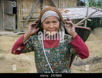 Raga, Arunachal Pradesh, India - 11 14 2010 : Rurale ritratto all'aperto di sorridente vecchia tribù Nyishi donna che porta il cestino conico tessuto con cinghia di testa Foto Stock