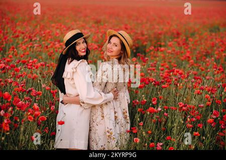 Due amiche in abiti e un cappello in un campo di papavero in estate al tramonto Foto Stock