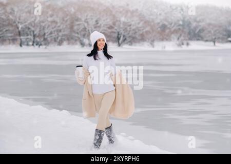 Una bella ragazza in un cardigan beige e un cappello bianco con un bicchiere di tè gode di un argine nevoso sul lago Foto Stock