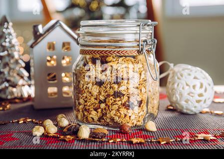 Concetto di regalo di Natale fatto in casa, granola arrosto fatta a mano con varie noci e uva passa in un bellissimo vaso di vetro. Offuscare l'albero di Natale e le luci. Foto Stock