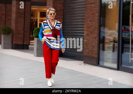 Donna alla moda che indossa abiti alla moda, pantaloni rossi, giacca blu, gilet a righe, scarpe bianche e occhiali da sole gialli. Modello con i capelli corti che camminano il Foto Stock